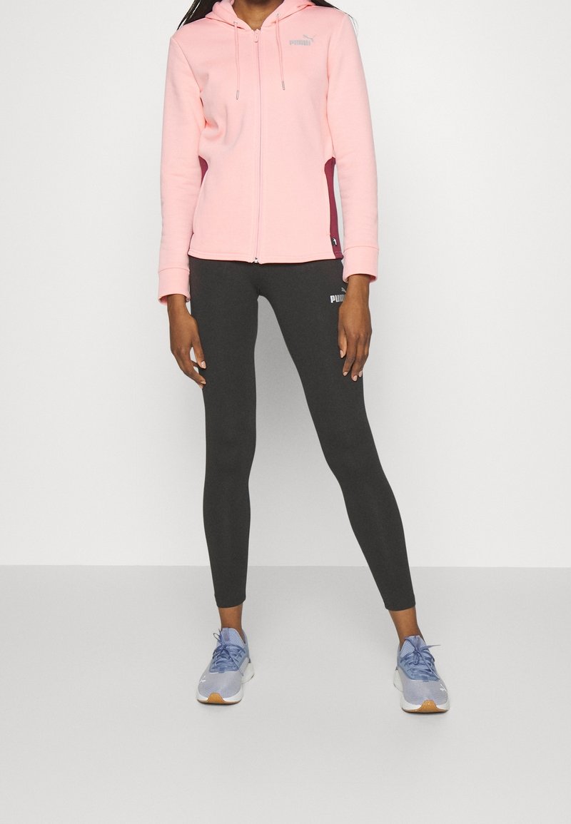 Femme debout portant un sweat à capuche rose zippé, des leggings Puma noirs et des chaussures de sport violet clair sur un fond blanc uni.