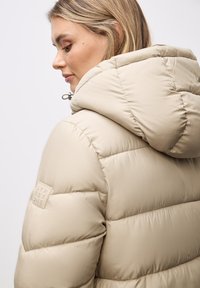 Giubbotto imbottito beige con cappuccio, caratterizzato da un design trapuntato e una toppa con il logo del marchio sulla manica. Texture liscia e forma strutturata.