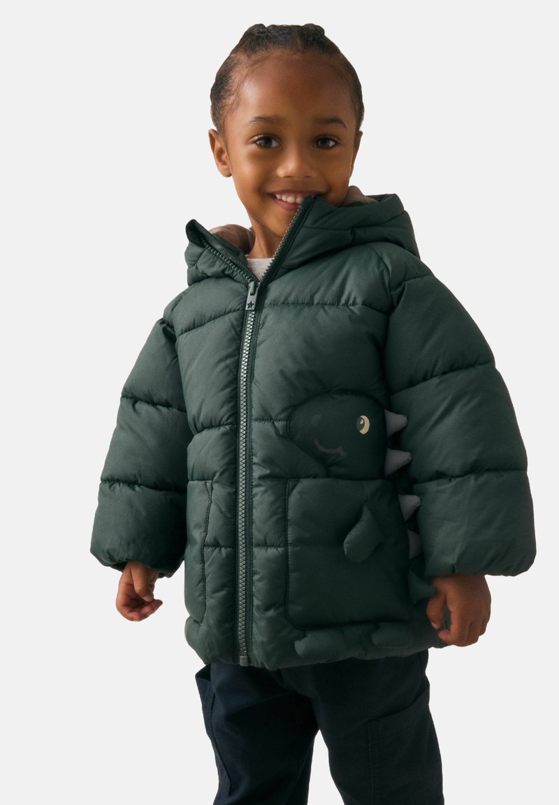 Jeune enfant souriant, portant une doudoune vert foncé avec une capuche et des détails de dinosaure sur la poche et les pointes des manches.