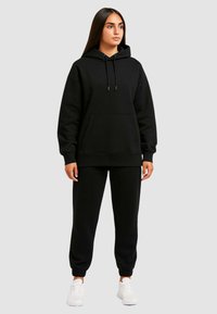 Completo felpa con cappuccio nera e pantaloni della tuta, caratterizzato da una tasca frontale, polsini a coste e coulisse. Indossato con sneakers bianche.