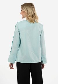 Blusa a righe blu chiaro con maniche lunghe, presenta dettagli con bottoni sulle spalle e una vestibilità ampia. Tessuto testurizzato con una leggera lucentezza.