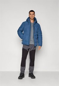 Mėlyna pūsta striukė su kapišonu, po ja pilka marškinėliai, derinami su juodomis ir pilkomis kelnėmis bei juodais batais. Lygi tekstūra.