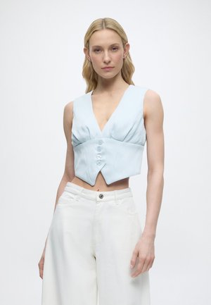 Ξανθιά γυναίκα με ανοιχτό μπλε αμάνικη κοντή μπλούζα με κουμπιά και λευκό παντελόνι με φαρδιά μπατζάκια και ψηλή μέση, στέκεται μπροστά σε απλό φόντο.