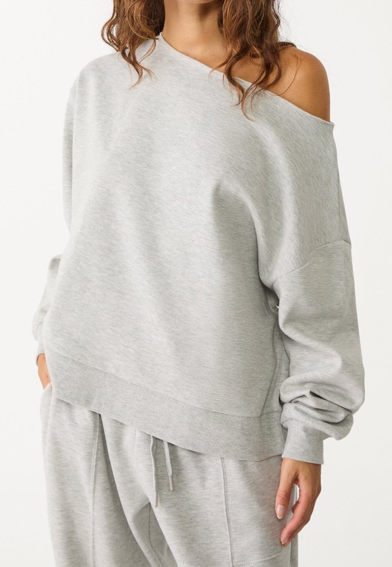 Femme portant un sweat-shirt oversize gris clair à épaules dénudées et un pantalon de jogging assorti avec taille à cordon, debout avec une main dans la poche.