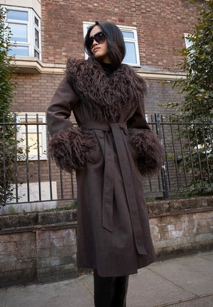 Brown long coat featuring a fuzzy collar and cuffs, belted waist, soft texture, and a loose fit. Styled with oversized sunglasses.