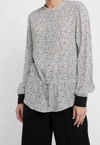 Blouse à manches longues en blanc et noir avec un motif floral, encolure ronde, ceinture élastique à la taille et poignets noirs. Tissu léger et texturé.
