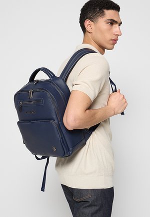 Mochila de cuero azul marino con acabado texturizado, múltiples compartimentos con cremallera y correas ajustables, llevada en un hombro.