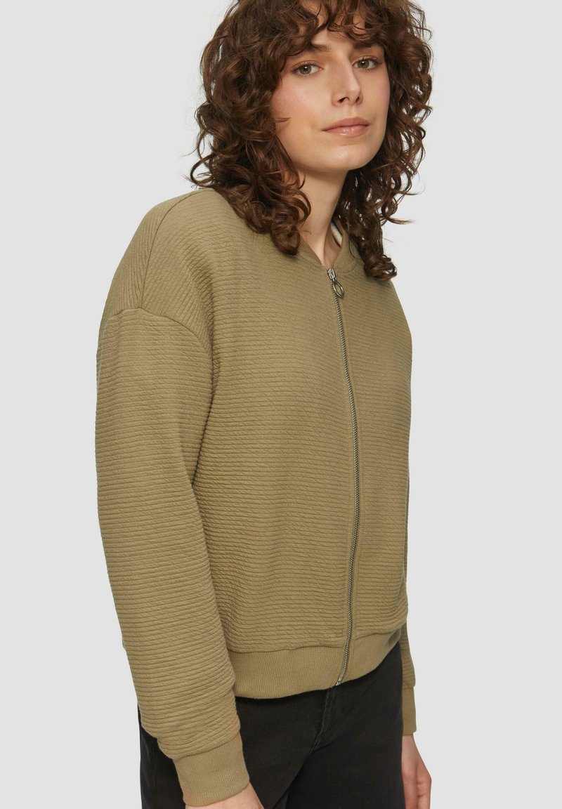 Olivovo zelená bunda na zips so štruktúrovanou rebrovanou tkaninou, vo voľnom strihu, s elastickými manžetami a lemom. Obsahuje kovový zips a minimalistický dizajn.