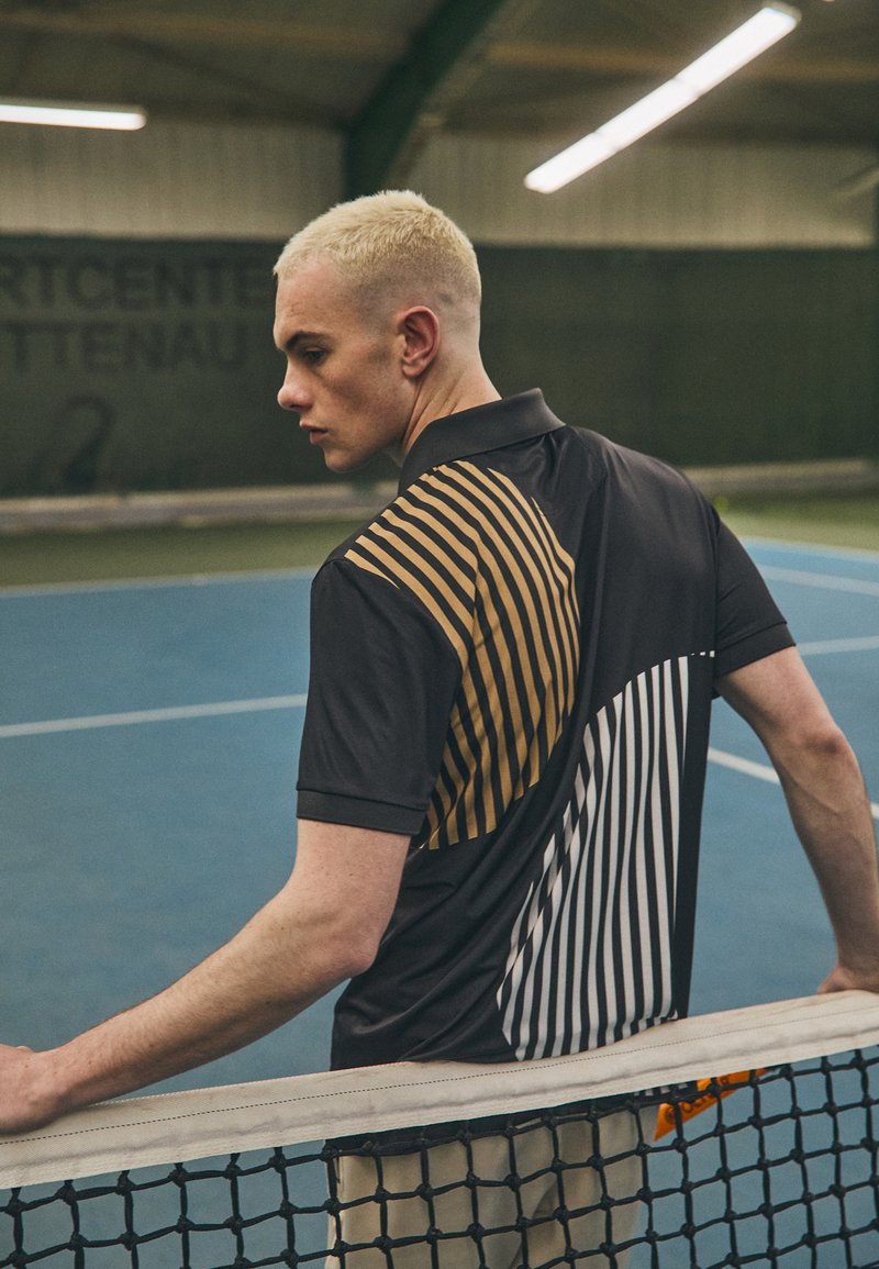 Black polo shirt featuring diagonal gold and white stripes on the back, slim fit, short sleeves, with tennis court backdrop.