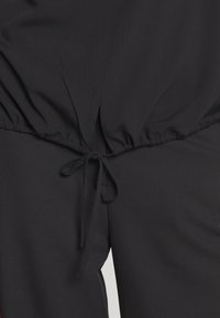 Haut en tissu noir avec une taille élastique et un cordon à nouer. Texture lisse et détail froncé à la taille.