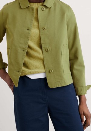 Man draagt een olijfgroene jas met knopen over een lichtgroene trui en donkerblauwe broek met hand in de zak.
