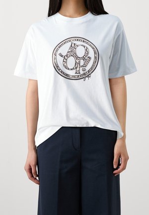 Person trägt ein weißes T-Shirt mit einem schwarzen und weißen, kreisförmigen "dp"-Logo in der Mitte, kombiniert mit lockeren schwarzen Hosen.