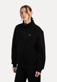 Mladá žena oblečená v černém pulovru na půl zip a černých kalhotách, stojící na světle šedém pozadí, směrem mírně doprava.