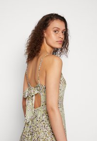 Blumendruckkleid aus leicht grünem Stoff, mit dünnen Trägern, einem Schleifenakzent am Rücken und einem offenen Rückendesign.