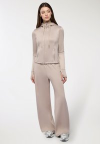 Felpa con zip color beige chiaro, dotata di cappuccio, maniche lunghe e aperture per le dita. Abbinata a pantaloni larghi coordinati. Entrambi hanno una texture liscia.