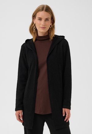 Femme blonde portant de grandes créoles dorées, un cardigan à capuche noir et un haut marron à manches longues, debout devant un fond clair uni.