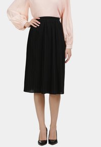 Vrouw in een zwarte geplooide midi-rok, een lichtroze blouse met lange mouwen en zwarte puntige high heels, staande met één hand op de heup.