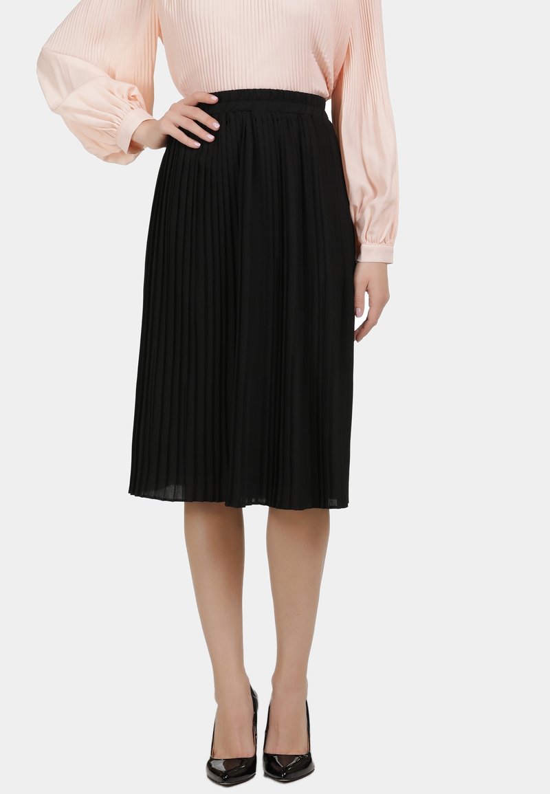 Vrouw in een zwarte geplooide midi-rok, een lichtroze blouse met lange mouwen en zwarte puntige high heels, staande met één hand op de heup.