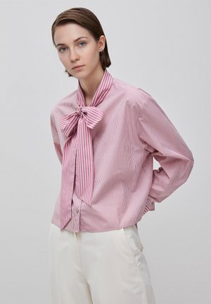 Frau mit kurzen braunen Haaren, die eine rosa gestreifte Bluse mit großer Schleife und weiße Hose trägt, vor einem schlichten hellen Hintergrund stehend.