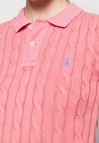 Różowa dziergana koszulka polo z fakturowanym wzorem warkocza, krótkim kołnierzykiem i trzema guzikami. Niebieskie logo w pobliżu klatki piersiowej.