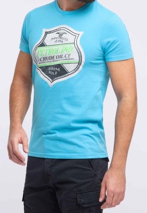 Mężczyzna noszący dopasowany jasnoniebieski t-shirt z czarno-zielonym, vintage’owym logo „Petrol Ind Crude Oil Co” oraz czarne szorty bojówki.