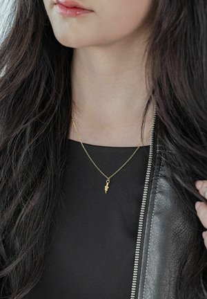 Junge Frau mit langen dunklen Haaren, die ein schwarzes Oberteil, eine Lederjacke und eine goldene Halskette mit einem kleinen Schlangenanhänger trägt.