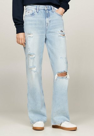 Jean déchiré bleu clair à jambes larges porté avec des baskets blanches et un haut sombre à manches longues, main dans la poche sur fond uni.