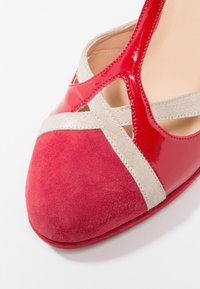 Chaussure en daim rouge et cuir verni avec des brides pailletées beiges qui se croisent sur l'avant-pied, montrant un gros plan du côté avant.