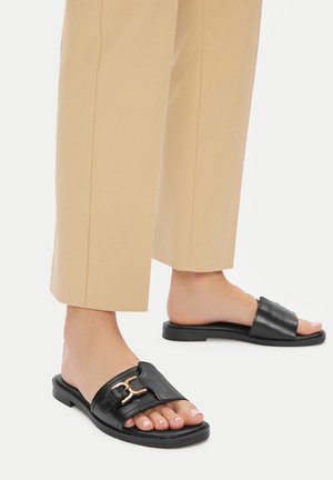 Czarne skórzane sandały na płaskiej podeszwie z szerokim paskiem i złotą sprzączką, zestawione z beżowymi spodniami o luźnym kroju.