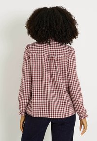 Camisa de cuadros rojos y blancos con mangas largas y puños fruncidos. Tiene un cierre de botones en la parte posterior y un ligero detalle de plisado. Textura de tela suave.