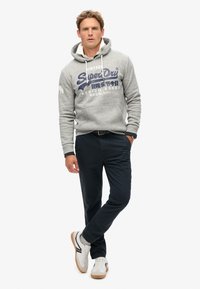Felpa grigia con design testuale frontale, lacci e polsini a costine, abbinata a pantaloni scuri e sneakers bianche con suola in gomma.