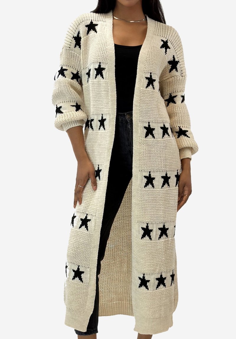 Langer cremefarbener Cardigan mit schwarzen Sternmustern, Strickstruktur, offenem Vorderteil und übergroßen Ärmeln. Getragen über einem schwarzen Oberteil und dunklen Jeans.