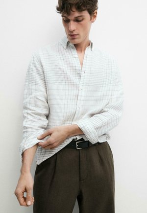 Giovane uomo che si arrotola la manica di una camicia bianca a quadri con bottoni, indossando pantaloni marrone scuro e cintura nera.