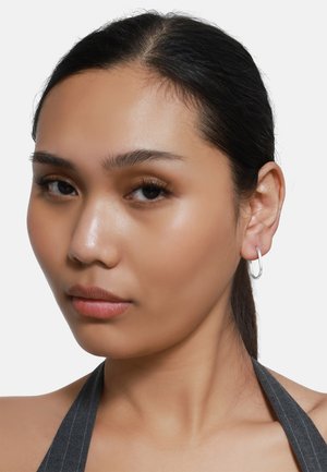 Les boucles d'oreilles créoles en argent présentent un design lisse et arrondi. Le modèle a les cheveux noirs raides et un teint neutre, mettant en valeur l'accessoire.