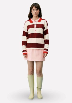 Femme portant un polo à manches longues rayé rouge et blanc, une mini-jupe rose et de grandes bottes en caoutchouc vert clair, debout sur un fond uni.