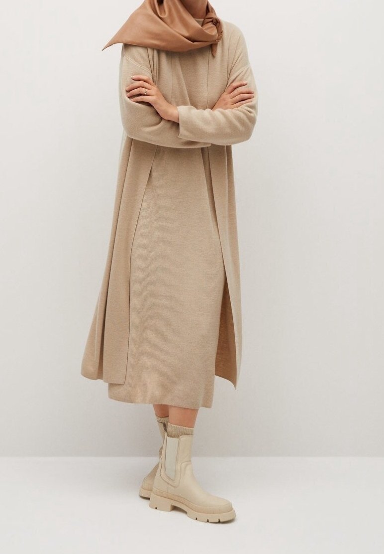 Femme portant une robe en maille côtelée beige, un long cardigan assorti, un foulard en satin couleur noisette sur la tête et des bottines crème épaisses avec des chaussettes, debout les bras croisés.