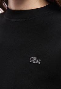 Κοντινό πλάνο στο γιακά ενός μαύρου πουκάμισου με ένα μικρό λευκό κεντημένο λογότυπο κροκοδείλου στο στήθος.