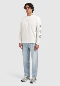 Wit shirt met lange mouwen met logo op de borst en grafische ontwerpen op de mouwen, gecombineerd met lichtblauwe jeans en metallic sneakers.