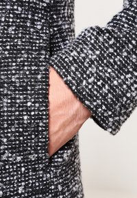 Black and white textured coat with a speckled pattern, featuring a visible pocket opening and a cuff detail, showcasing woven fabric.