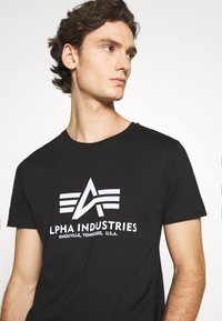 Sort bomulds-t-shirt med et hvidt "ALPHA INDUSTRIES" logo med et emblem, rund hals, korte ærmer og en klassisk pasform.