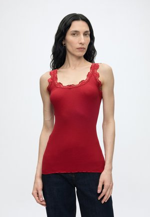 Femme aux longs cheveux foncés portant un haut côtelé rouge sans manches avec bordure en dentelle et un jean foncé, debout devant un fond clair uni.