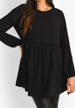 Femme portant un haut tunique noir ample à manches longues avec taille froncée, associé à des leggings noirs et un collier doré, sur fond blanc.