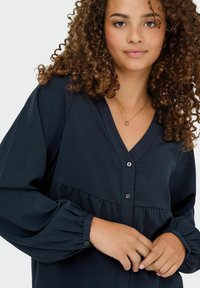 Navy blouse with a V-neck, long puff sleeves, and buttons down the front. The fabric has a textured finish and a gathered waistline.
