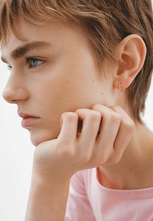 Pendiente de aro dorado con pequeños acentos colgantes, llevado en la oreja de una persona con cabello corto y piel clara, apoyando su mano en el mentón.