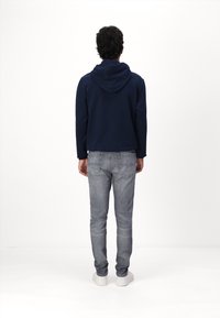Muž stojící zády, oblečený v tmavě modré mikině s kapucí, šedých slim-fit džínách a bílých teniskách na čistém bílém pozadí.