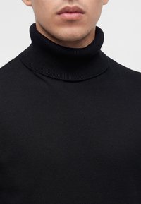 Gros plan d'une personne portant un pull à col roulé côtelé noir, montrant le cou et la partie inférieure du visage avec des lèvres neutres.