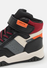 Basket haute avec des panneaux noirs, gris, crème, rouge et orange, une sangle Velcro avec le logo Geox, et des lacets noirs à rayures rouges.