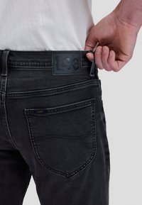 Hand stellt Gürtelschlaufe an schwarzen Lee-Jeans ein, getragen mit weißem Hemd, zeigt Gesäßtasche und Markenlabel am Bund.