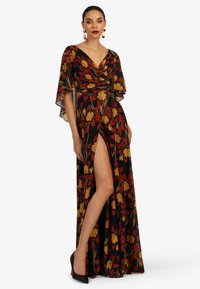 Maxikleid aus schwarzem Stoff mit rotem und gelbem Blumenmuster, Wickeloberteil, schwingenden Ärmeln und einem Schlitz bis zum Oberschenkel. Schwarze spitze Absätze.