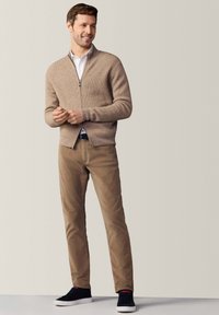 Maglione beige con zip e texture a costine, abbinato a pantaloni beige e sneakers nere con suola bianca. Design semplice e silhouette aderente.
